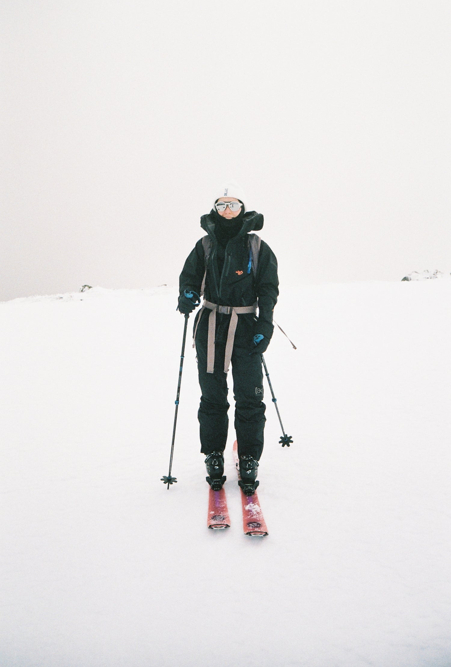 An image of owner, Abigail Burgess ski touring in white out conditions. She is wearing pink skis and all black outerwear with sunglasses and a white beanie.
