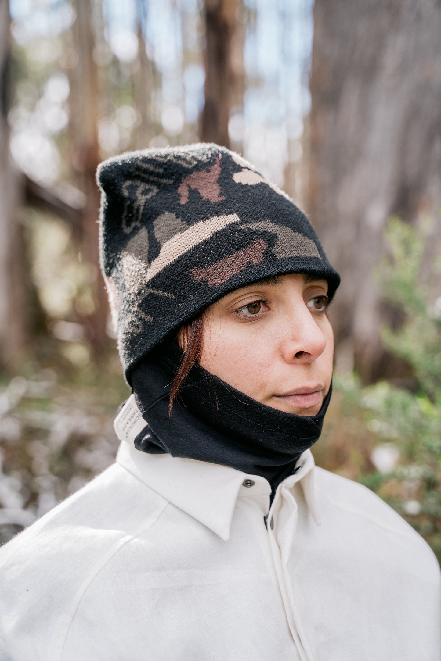 A woman pictured wearing a black neck tube and a camo beanie, facing slightly side onto the camera