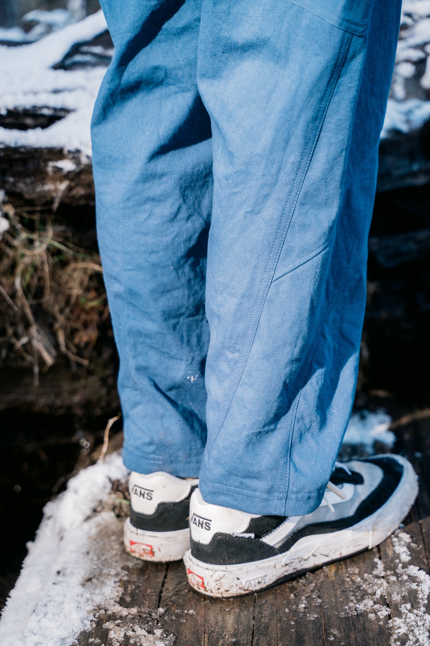 An image of the bottom of blue pants. A man is wearing them standing on a log with some snow.