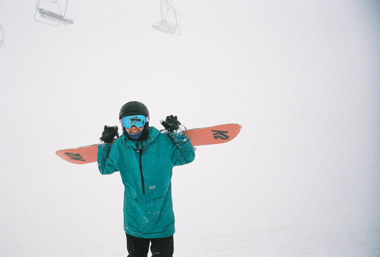 A man holding a snowboard over his shoulders walking up a hill.  It is misty and he is wearing a blue Schusser Threads anorak, black helmet and pants and blue goggles.