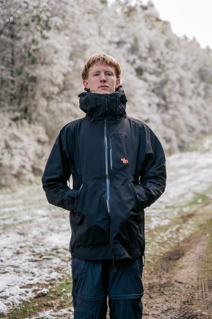 A man wearing a black jacket and black pants, standing in a snowy landscape facing the camera.