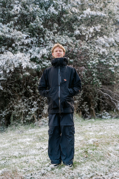 A man wearing a black jacket and black pants, standing in a snowy landscape facing the camera.