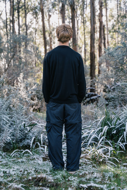 A man standing in the bush with a light covering of snow wearing a black long sleeve shirt and black pants