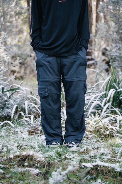 The bottom half of a man wearing a black shirt and pants, standing in some grass that has a light dusting of snow.