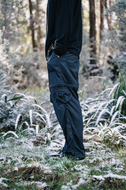 The bottom half of a man standing in some grass with a light covering of snow.  The man is wearing a black shirt and pants.
