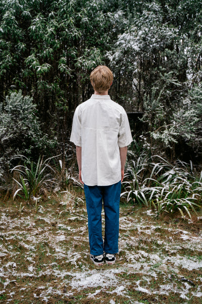 A man in a white shirt and blue pants standing in snow covered bush facing away from the camera.