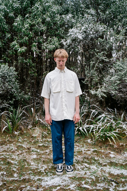 A man in a white shirt and blue pants standing in snow covered bush facing towards the camera. 