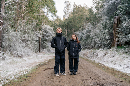 A man and a woman wearing a black jacket and black pants, standing in a snowy landscape facing the camera.