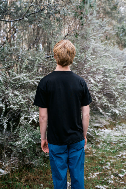A man standing in a snowy landscape wearing a black tee and blue pants facing awa from the camera.