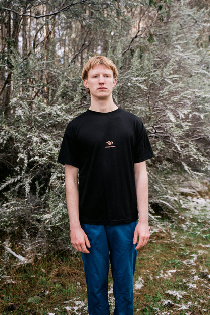 A man standing in a snowy landscape wearing a black tee and blue pants facing towards the camera.