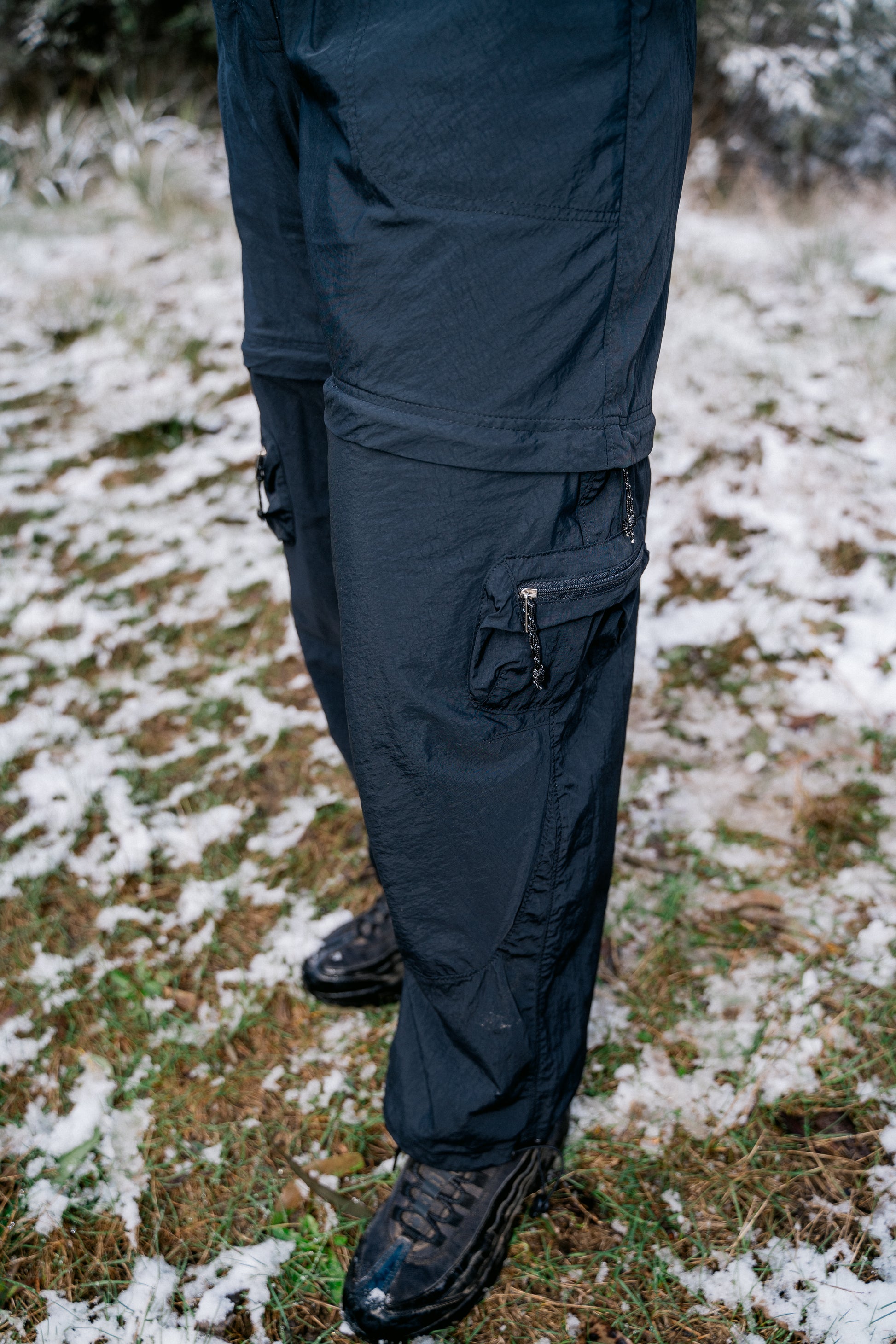 A womens legs wearing long black pants that can zip off. She is standing in some grass with a light dusting of snow.