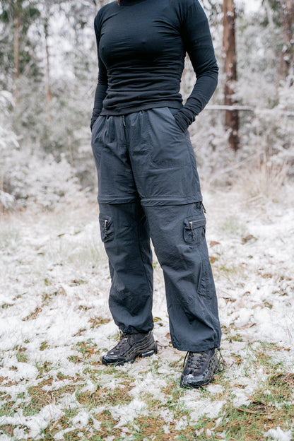A women (face not pictured) standing slightly side on to the camera wearing a black shirt and pants in a landscape that has a light covering of snow.