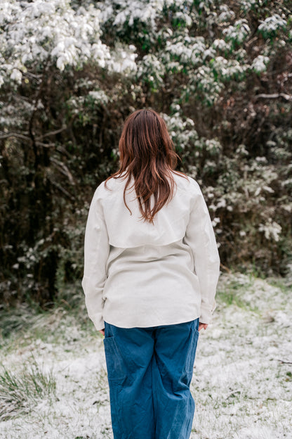 A woman standing in a white shirt and blue pants in the bush with a light dusting of snow facing away from the camera.