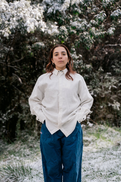 A woman standing in a white shirt and blue pants in the bush with a light dusting of snow facing the camera.