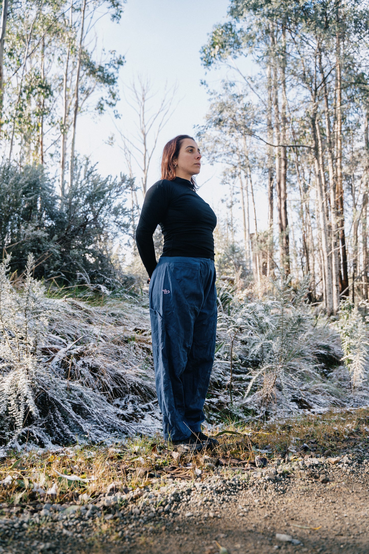  A full photo of a woman wearing blue pants and a black shirt standing in the grass with a light dusting of snow facing to the side.