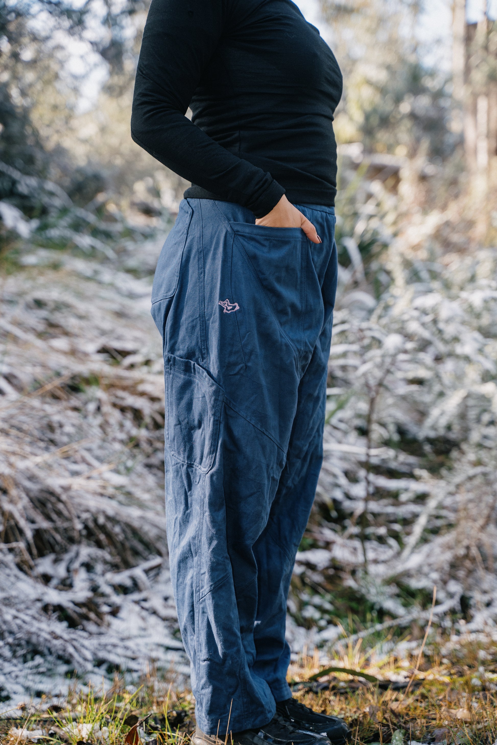 A woman wearing blue pants and a black shirt standing in the grass with a light dusting of snow facing to the side.
