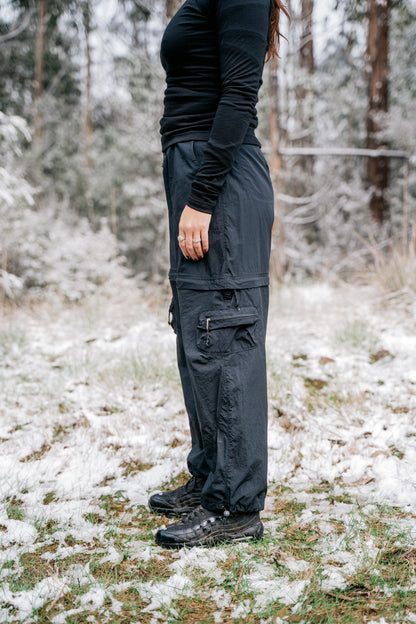 a woman standing side on wearing all black in a grassy glade with a light dusting of snow