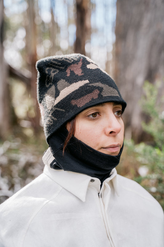 A woman pictured wearing a black neck tube and a camo beanie, facing slightly side onto the camera
