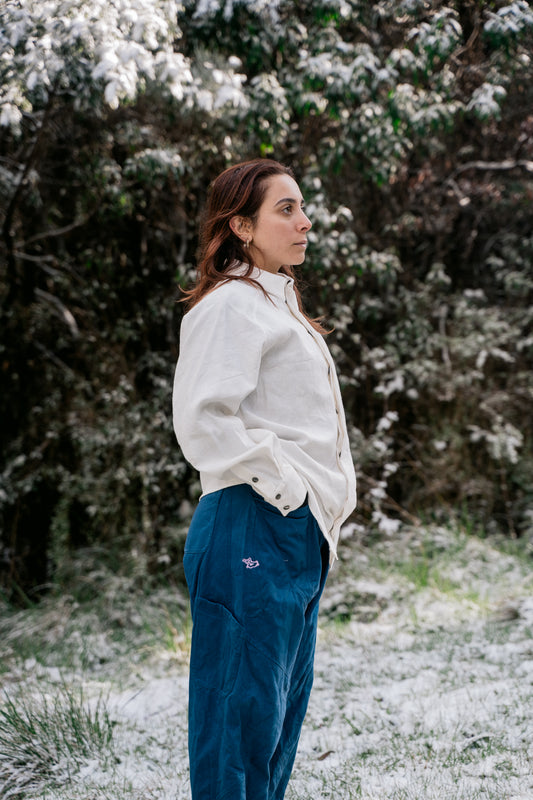 A woman standing in a white shirt and blue pants in the bush with a light dusting of snow standing side on to the camera.