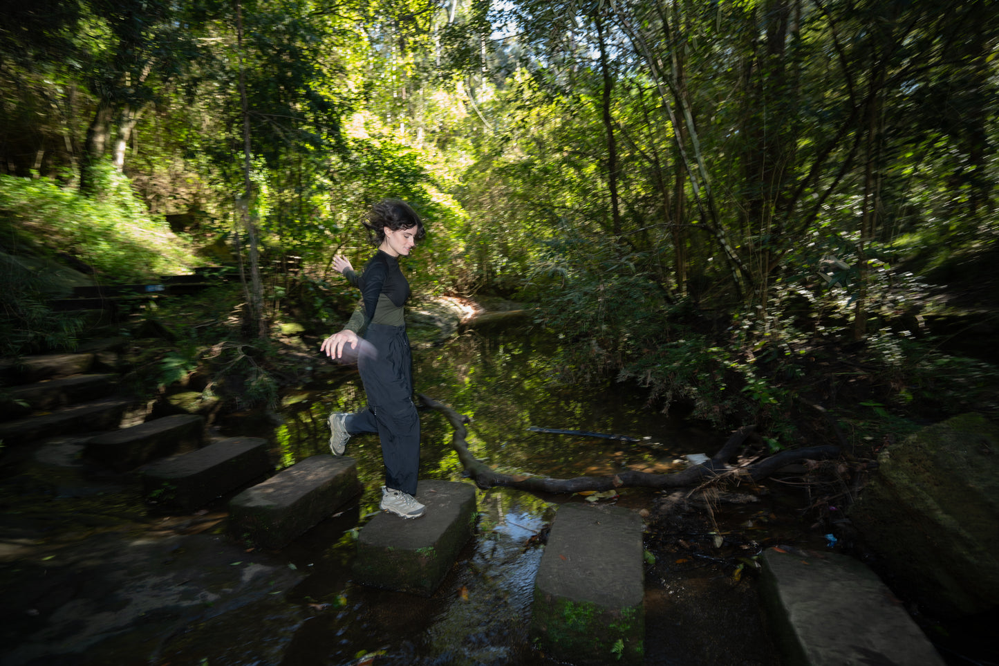 A woman rockhopping across a creek in black pants and a black and olive shirt.