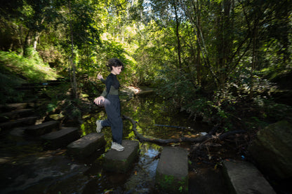 A woman rockhopping across a creek in black pants and a black and olive shirt.