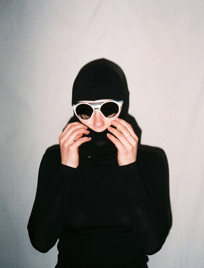 A front on photo of a girl wearing white sunglasses and a black balaclava with her hands brought up to her face