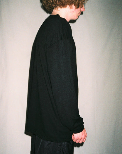 A man standing in front of a white background wearing a black long sleeve shirt side on.