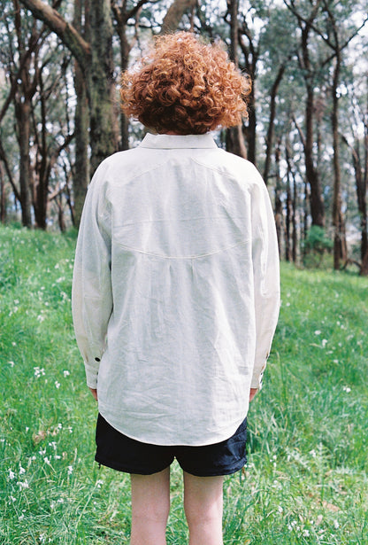 An image of a man standing in some lush grass with trees in a white long sleeve collared shirt