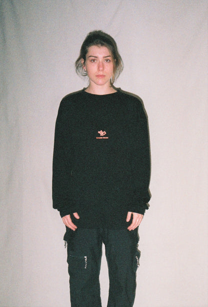 A girl standing in front of a white background wearing a black merino shirt with a red logo